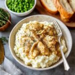 Bowl of chicken and gravy over creamy mashed potatoes with peas and bread on the side